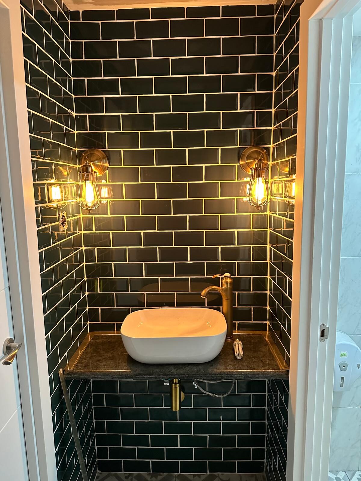 Zona de lavabo terminada con azulejo metro negro, grifería dorada y apliques industriales